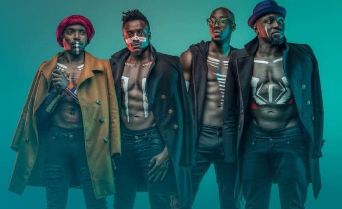 67df943dc8c4f6001dd22265.jpg Four men in coats from the Afro-Pop band Sauti Sol stand confidently against a teal background. They have painted designs on their bare chests and wear hats or a headwrap, embodying the vibrant spirit of Kenya.