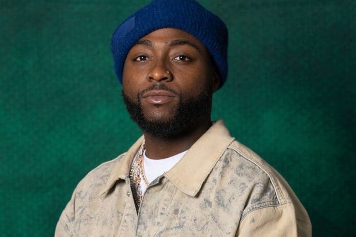 A man with a beard wearing a blue knit hat, beige jacket, and white shirt stands in front of a green textured background, exuding cool Afrobeats vibes reminiscent of Davido.