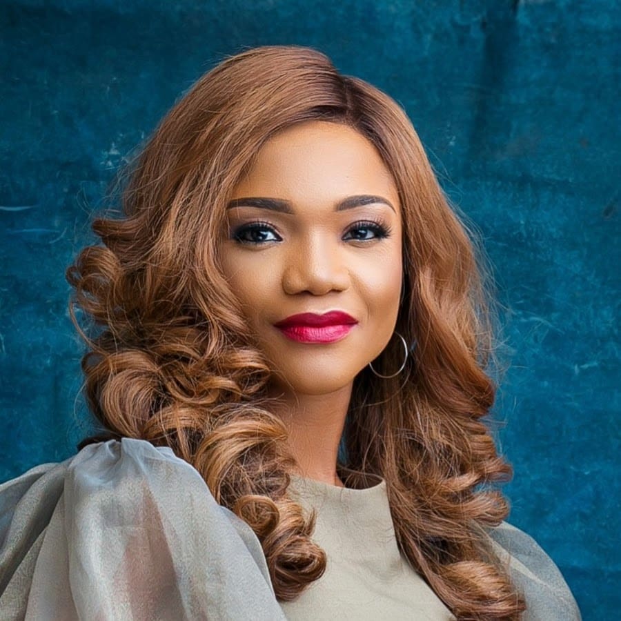 Smiling person with long, curly brown hair and red lipstick, wearing silver hoop earrings and a sheer sleeve top, against a blue textured background.
