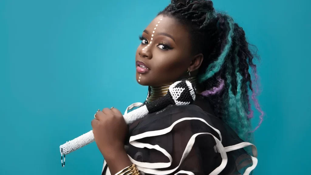 A woman with beaded jewelry and face paint holds a decorated microphone against a turquoise background, wearing a black outfit with white trim.
