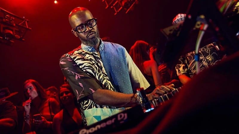 A DJ wearing glasses and a patterned shirt, inspired by Black Coffee, performs on stage using a Pioneer DJ controller, with a crowd and colorful lights in the background.
