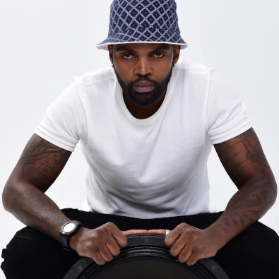 Man in a white t-shirt and patterned hat sits on a speaker, holding its edges.
