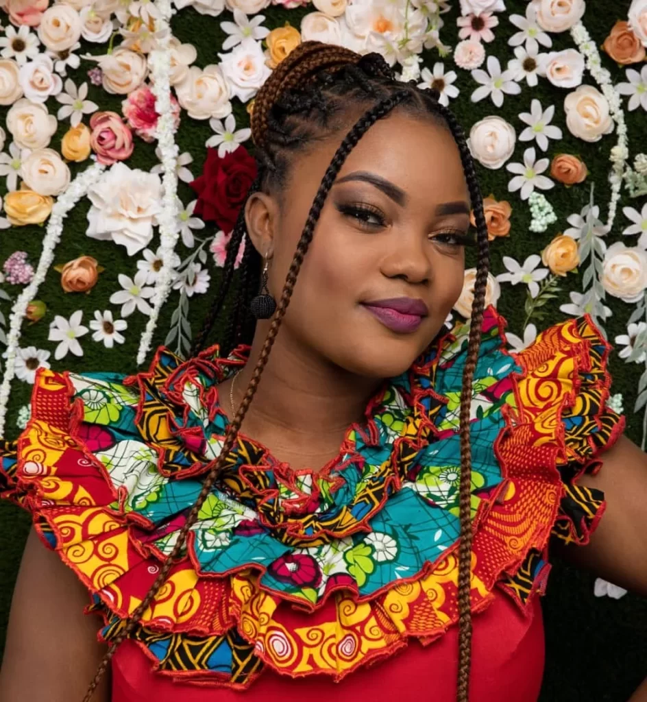 A woman with braided hair and bright makeup wears a colorful ruffled dress, standing in front of a wall decorated with various flowers.