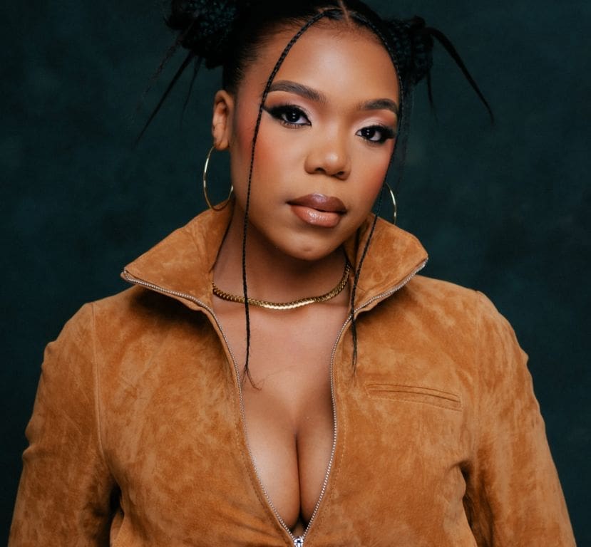 A woman with braided hair poses against a dark background wearing a tan suede jacket, hoop earrings, and a gold necklace.
