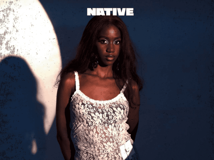 IMG_5619.png Person in a white lace top stands against a textured wall, partially illuminated by bright light, with the word "NATIVE" above them. It's Maya Amalo gracing the cover of NativeMag with an enigmatic profile.
