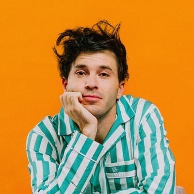 A person with messy hair, reminiscent of Jeremy Loops' carefree style, wearing a green and white striped shirt, rests their chin on their hand against an orange background.