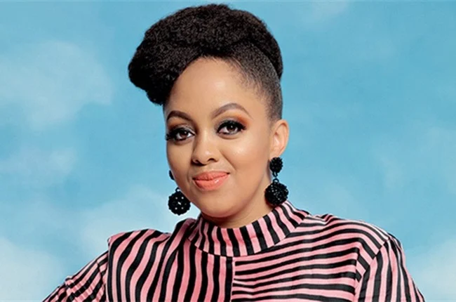 A woman with an updo hairstyle, wearing black earrings and a pink and black striped top, poses in front of a plain blue background, radiating the spirit of joyous celebration.