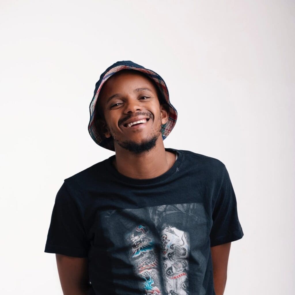 A smiling person, styled like Kabza De Small, dons a bucket hat and a graphic T-shirt, posed effortlessly against a plain background.