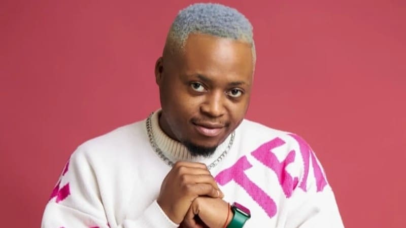 A person with short, dyed hair, wearing a white sweater with pink text, poses against a red background with hands clasped.