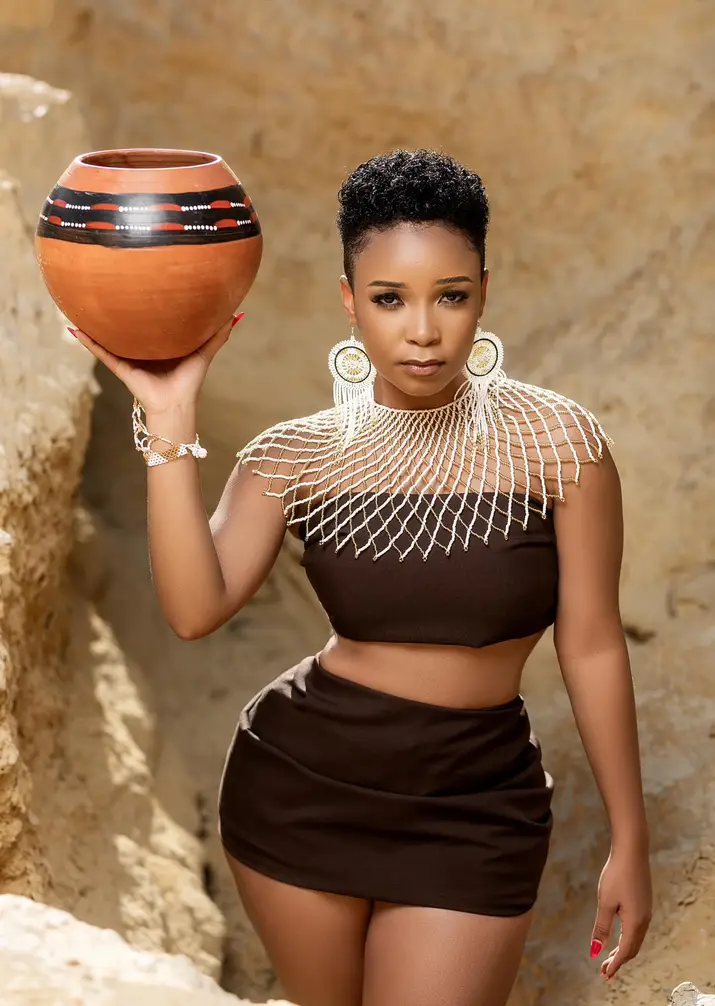 Kharishma wearing a brown outfit and beaded accessories holds a decorative pot on one shoulder, standing against a rocky background.