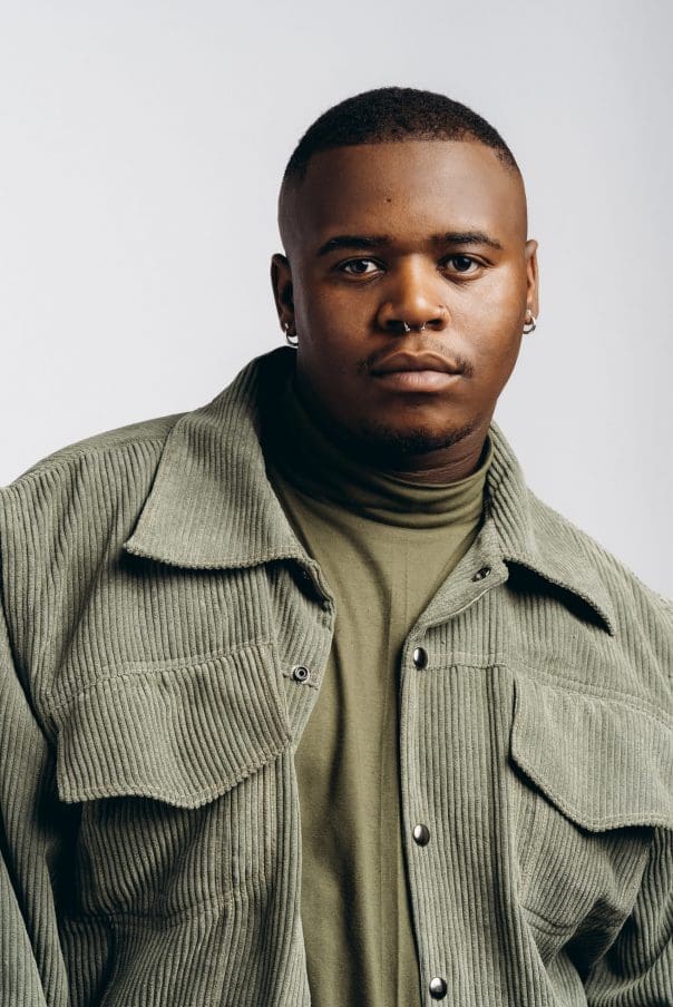 A person with short hair wearing a green turtleneck and corduroy jacket against a light background.