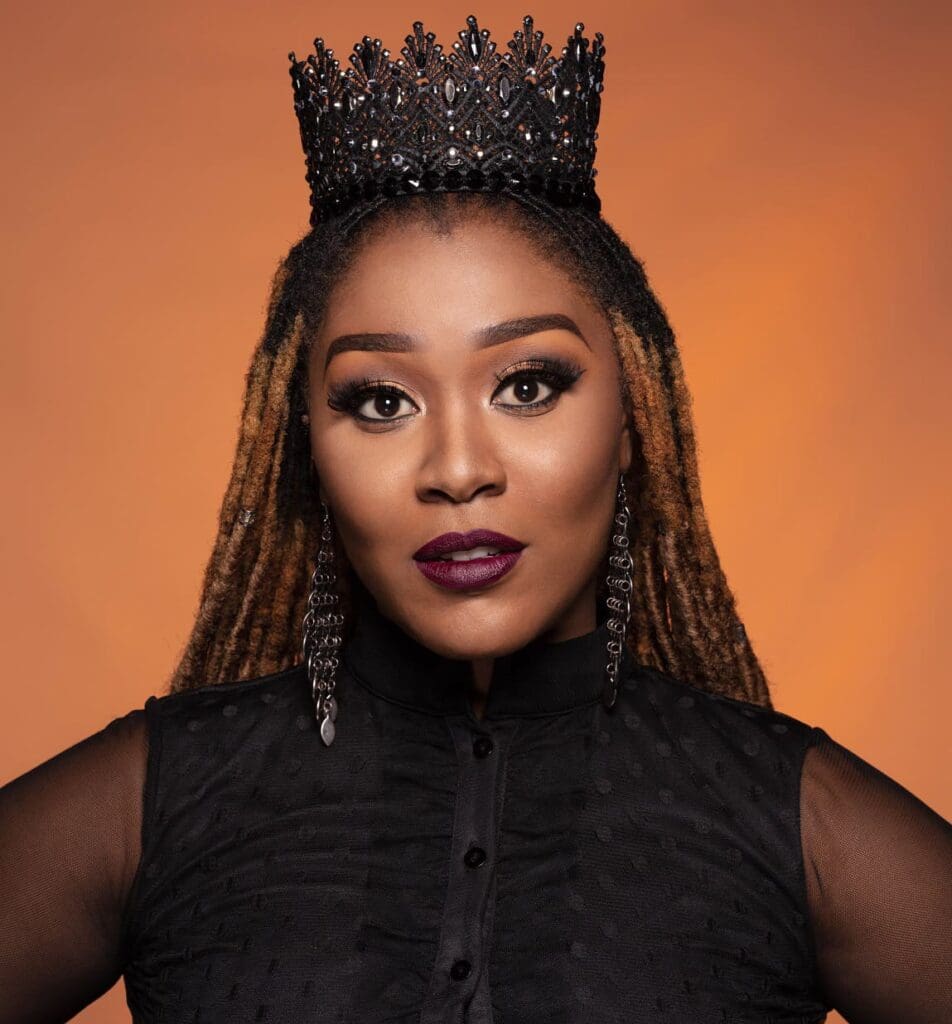 A woman with long, highlighted dreadlocks wears a black beaded crown, bold makeup, long earrings, and a black sheer-sleeved top against an orange background.