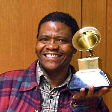 Ladysmith Black Mambazo in a plaid shirt holds a Grammy award in one hand, standing in front of a wooden background.