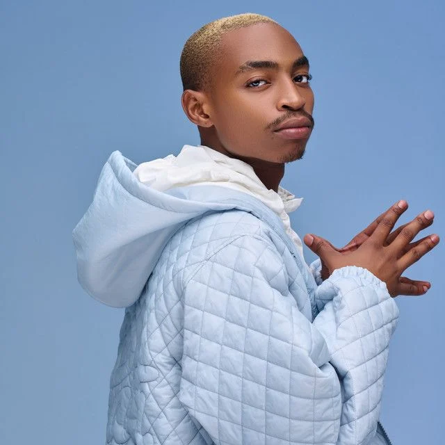 Person with blond hair wearing a light blue quilted jacket over a white hoodie, posing against a blue background.