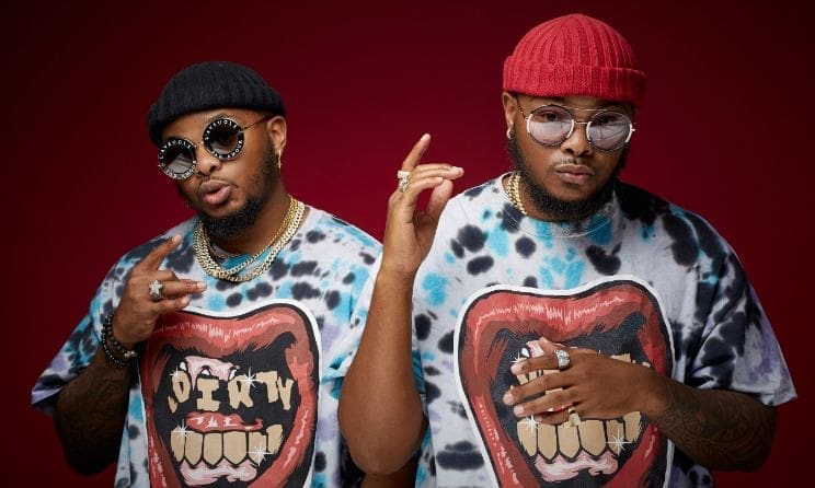 Two men, channeling a Major League DJz vibe, sport sunglasses, knitted beanies, and tie-dye shirts with graphic designs as they pose against a dark red background.