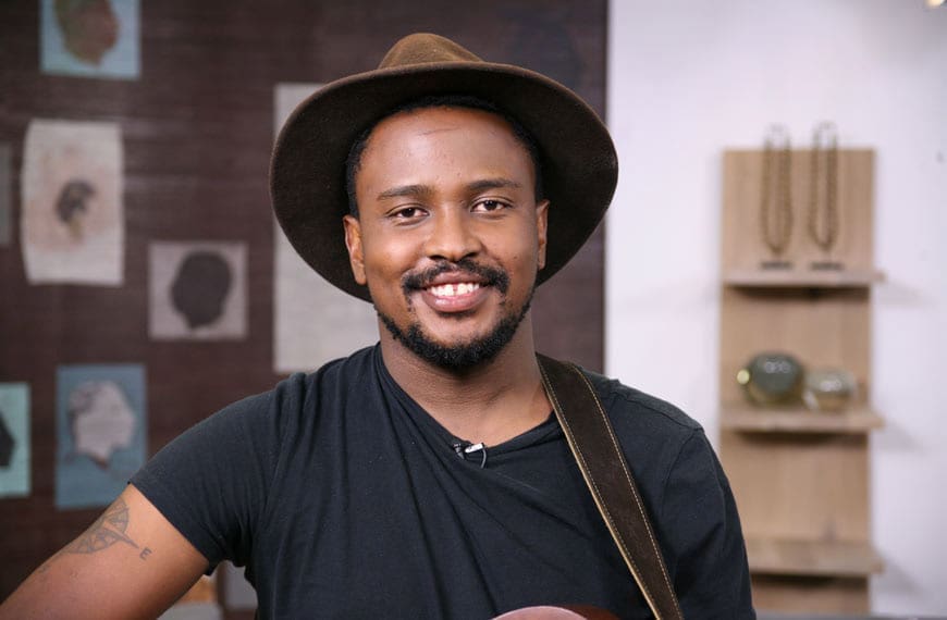 A man wearing a brown hat and black t-shirt smiles at the camera, with framed artwork and jewelry displays in the background.