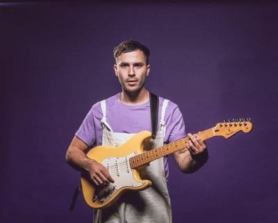Person wearing a lavender shirt and light overalls plays an electric guitar against a plain purple background.