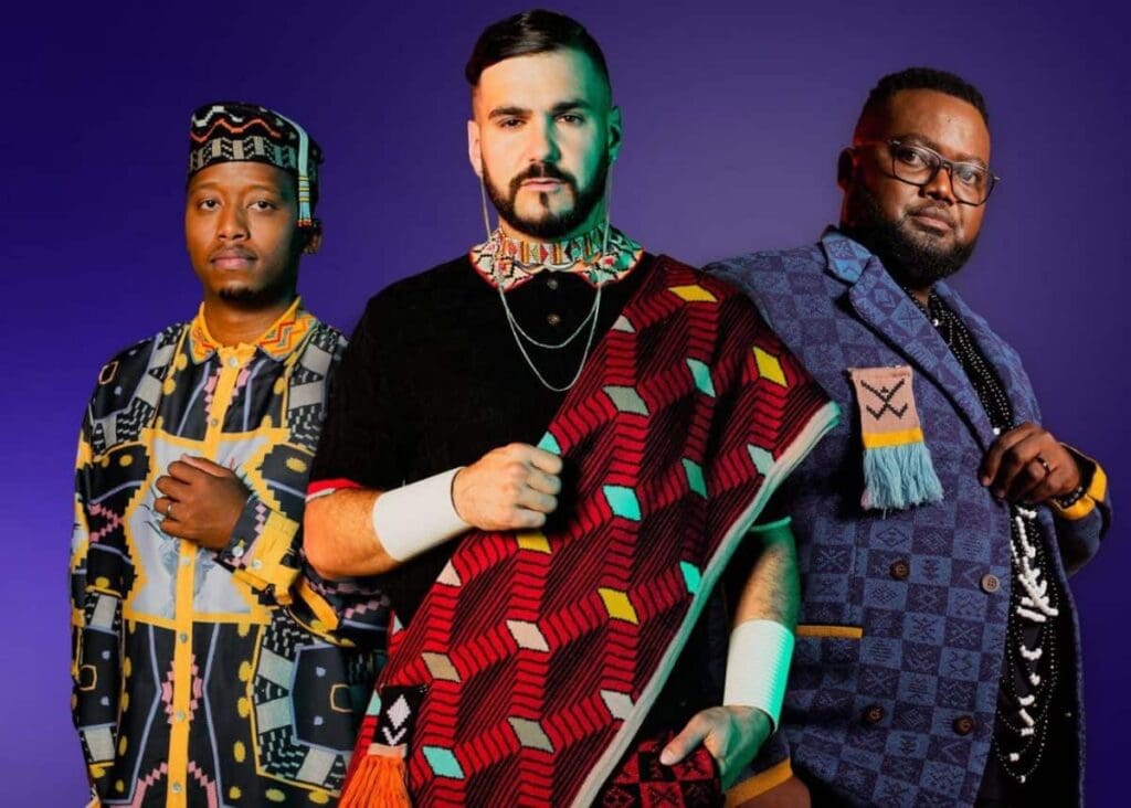 Three men stand against a purple background, wearing colorful, patterned traditional and modern clothing and accessories, facing the camera with neutral expressions.