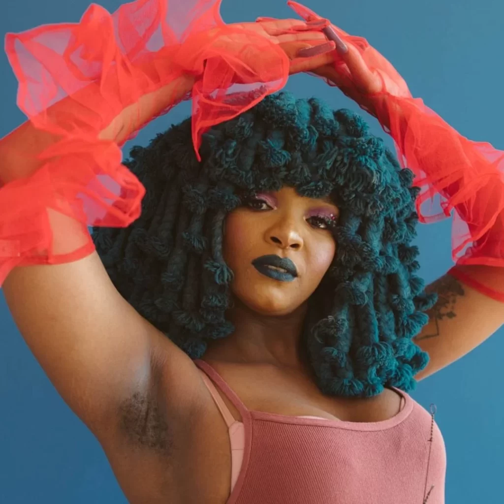 Moonchild Sanelly poses against a blue background with their arms raised. Sporting a large teal wig, red mesh gloves, and a pink top, this vibrant look is unmistakably Moonchild Sanelly, the acclaimed South African musician and Gqom artist known for her unique style and energetic performances.