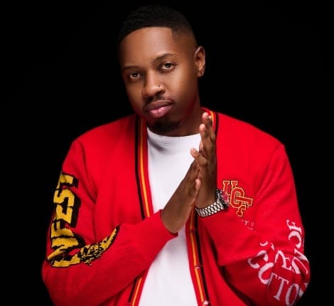 Man wearing a red jacket with text, clasping hands together against a black background.