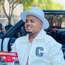 Mthandeni SK, in a white hat and jacket, stands beside an open car door outdoors, with trees and buildings softly framing the background.