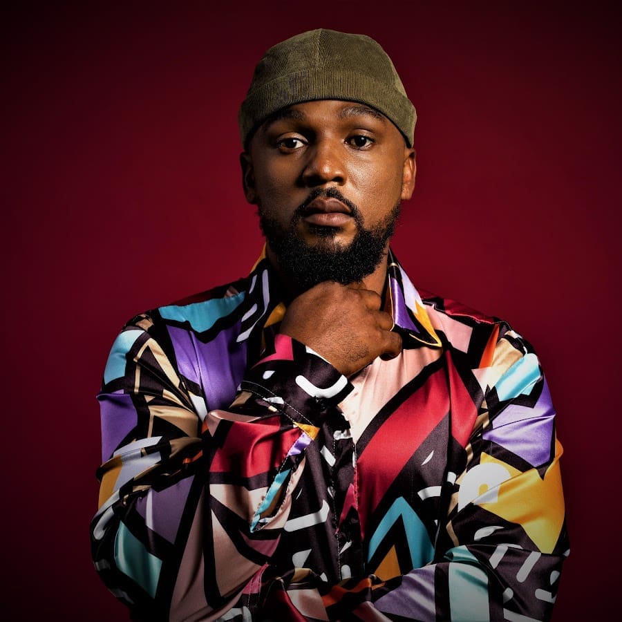 A man wearing a colorful geometric-patterned shirt and olive green cap poses against a dark red background, with one hand touching his neck.
