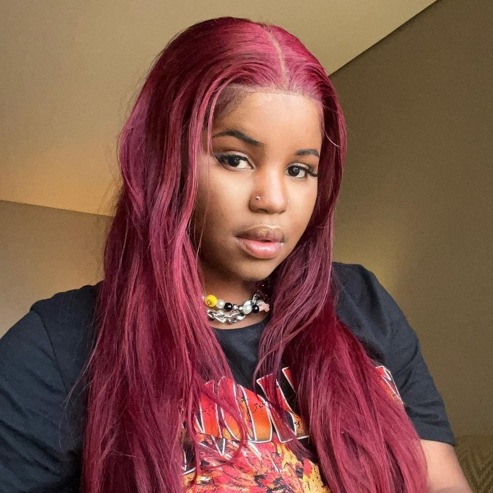 Person with long, straight, dark pink hair and a nose piercing, wearing a black graphic T-shirt and a chunky necklace, poses indoors against a plain background.