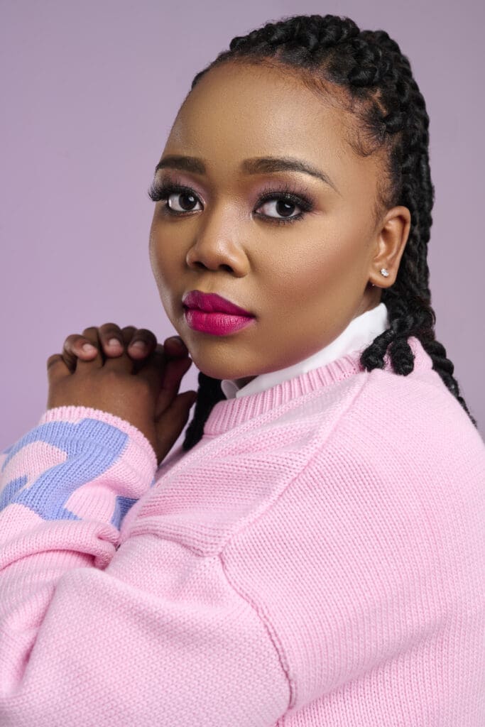 Nontokozo Mkhize with braided hair and pink lipstick wears a light pink sweater, looking at the camera with her hands clasped near her face against a light purple background.