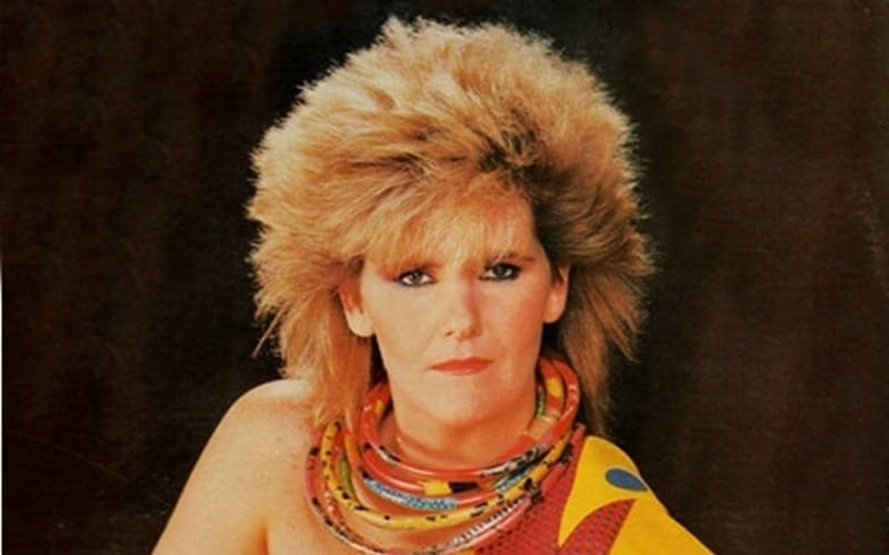 A person with voluminous, teased blonde hair and layered necklaces poses against a dark background, wearing a colorful garment draped over one shoulder.