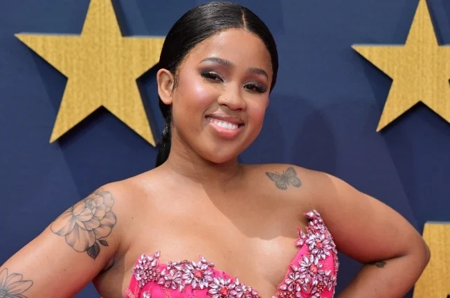 Person smiling in a pink dress with floral embellishments, standing in front of a backdrop with large stars.