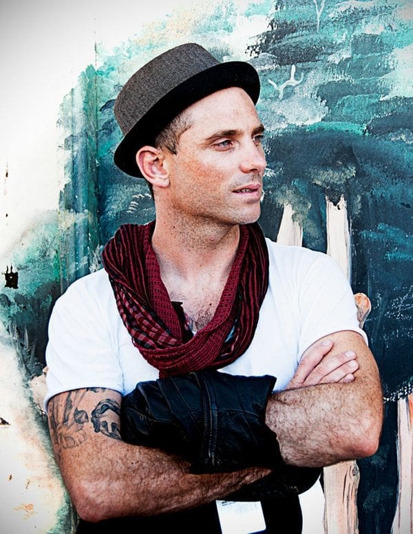 Man in a hat with a red scarf and tattoos stands with arms crossed against a painted backdrop.