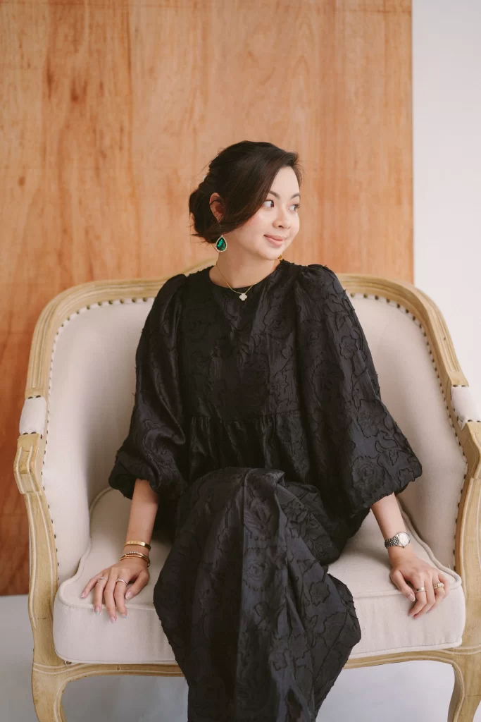 Woman in a black dress sits on a beige armchair against a wooden backdrop.