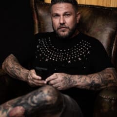 A person with tattoos sits in a leather armchair, holding a smartphone and looking at the camera. They wear a black shirt with a studded design, exuding the edgy vibe of a Prime Circle concert.