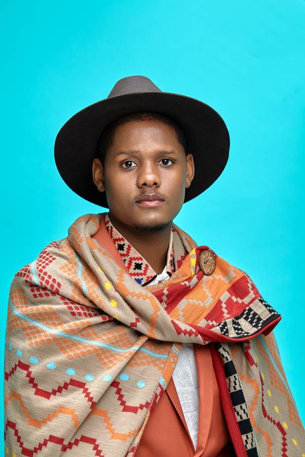 A person wearing a wide-brimmed hat and a patterned, colorful shawl over a suit jacket poses against a solid turquoise background.
