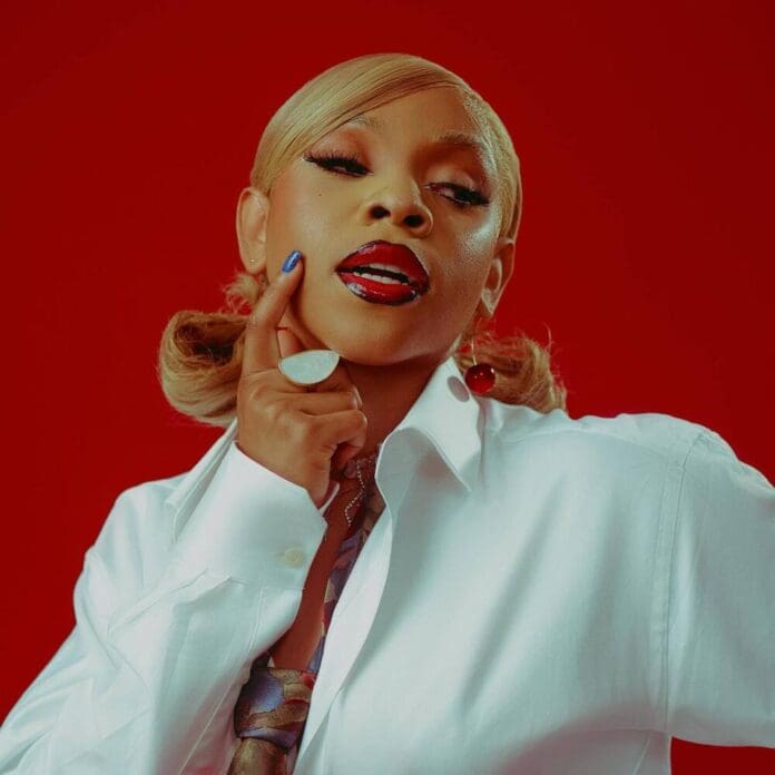 SaveClip.App_461228746_18463287019046202_2894544294886272957_n-1.jpg Person with blond hair poses against a red background, wearing a white shirt and touching their chin, channeling the vibrant energy of Afrobeats.