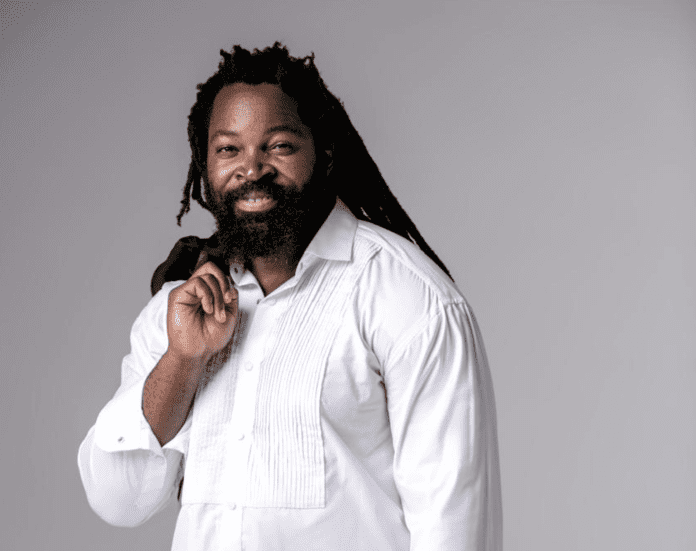 Screenshot-2025-04-11-at-20.33.33.png Man with a beard and dreadlocks smiling, wearing a white shirt, standing against a gray background—reminiscent of Big Zulu on the cover of his new album "Icala LeMpumelelo.