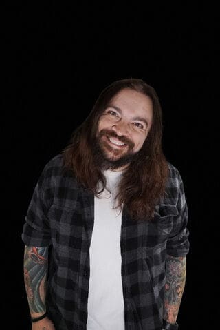 Man with long hair and tattoos smiles, wearing a plaid shirt over a white T-shirt, against a black background.
