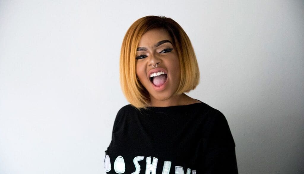 Woman with a blonde bob haircut wearing a black shirt stands against a plain white background, mouth open as if singing or shouting.