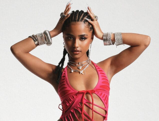 A woman poses with her hands on her head, wearing a pink cut-out top, multiple silver bracelets, and layered necklaces.