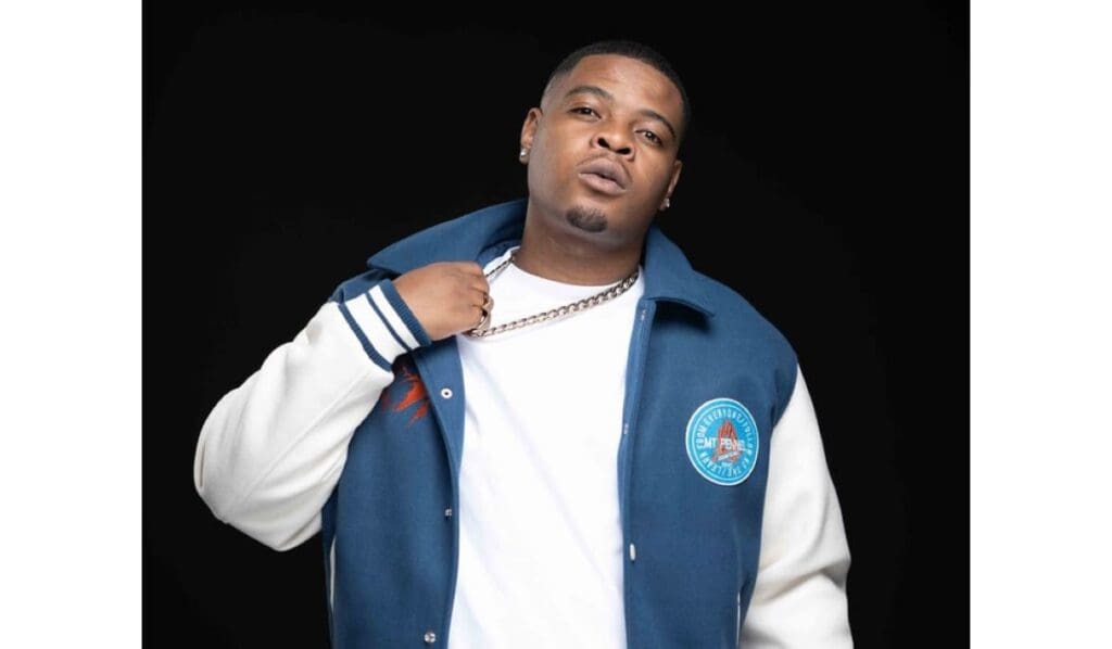 A man in a blue and white jacket holds a chain around his neck against a black background.