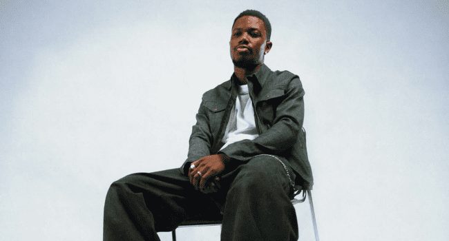 Tyson-.png A person, reminiscent of Tyson Sybateli awaiting a music release, sits on a chair against a plain light background, dressed in a dark jacket, white shirt, and dark pants, looking slightly to the side.