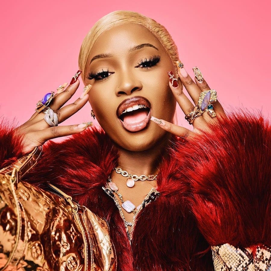 A person with platinum blonde hair and elaborate makeup, reminiscent of Uncle Waffles' vibrant style, poses against a pink background, adorned with multiple rings and a furry red coat.