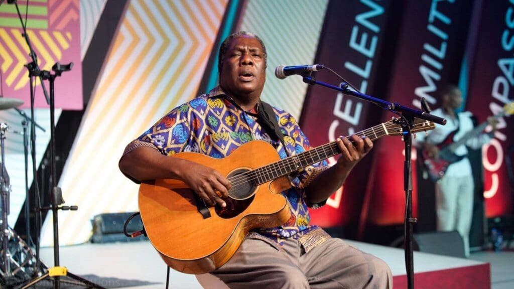 A man sits onstage playing an acoustic guitar and singing into a microphone, with a colorful backdrop and other musicians in the background.
