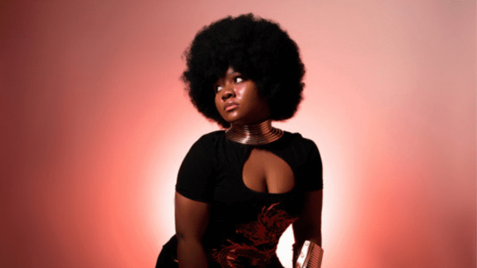 Wiziboy_20250401_200732_0000.png Mary-Lue, with her striking afro hairstyle, exudes passion in a black dress adorned with gold accessories. She stands poised against a captivating pink and red gradient background that radiates warmth and intensity.