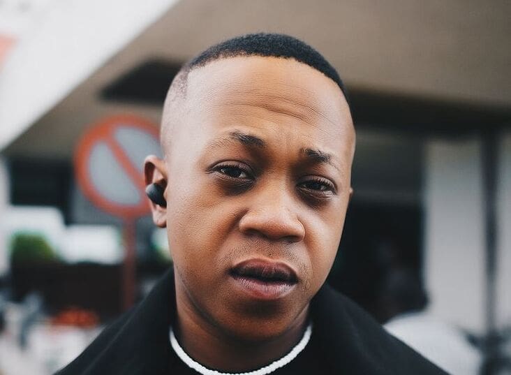 A person, reminiscent of a Young Stunna vibe, wears a black coat and earpiece while standing outdoors with a no-parking sign in the background.