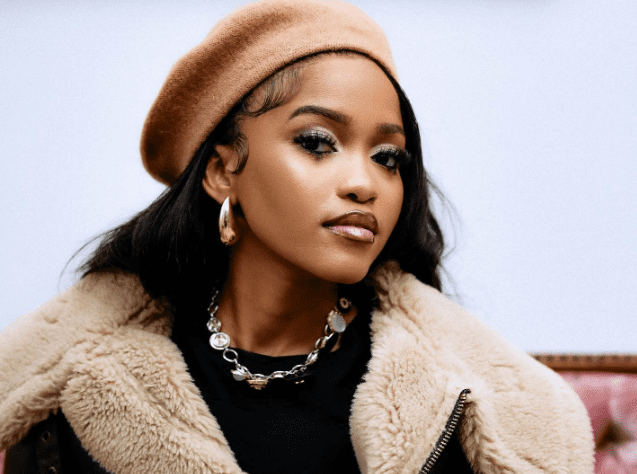 A woman wearing a tan beret, gold hoop earrings, a chunky necklace, and a shearling-lined coat sits against a plain background, looking confidently at the camera.