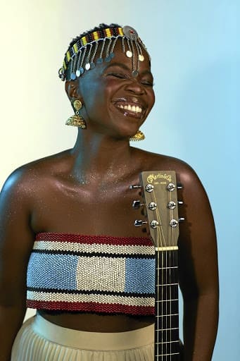 Woman wearing beaded headpiece and earrings, smiling with eyes closed, stands beside an acoustic guitar and wears a beaded bandeau top.