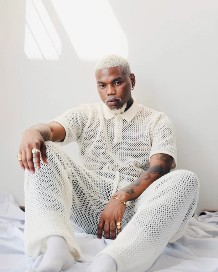 halo.webp.webp Person with short blonde hair wearing a white mesh outfit, sitting on a white blanket in a well-lit room, tattoos visible on arms. They radiate the edgy vibe of Phresha's new single, blending art and attitude seamlessly into their look.