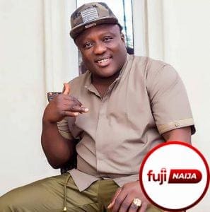 Man sitting in a chair, wearing a brown shirt and camouflage cap, making a hand gesture. A "fuji NAiJA" logo is in the bottom right corner.