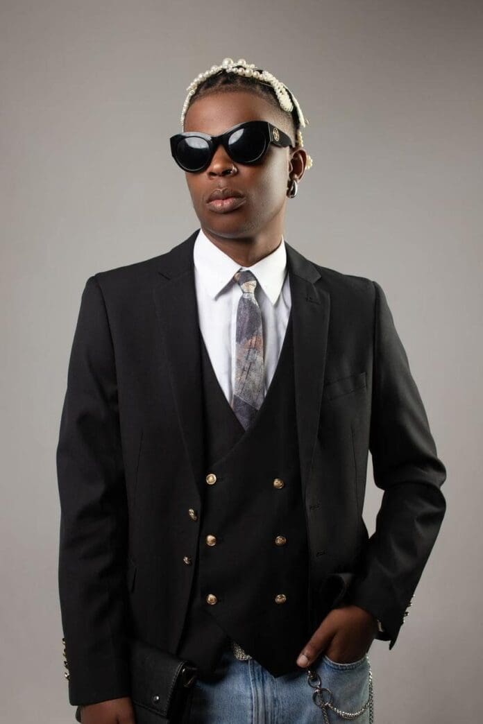 unnamed-file.jpeg Person wearing a black suit with a vest, tie, and sunglasses stands against a plain background. They have braided hair and are holding a black clutch, exuding elegance reminiscent of Aubrey Qwana's style, subtly reflecting their Zulu heritage.